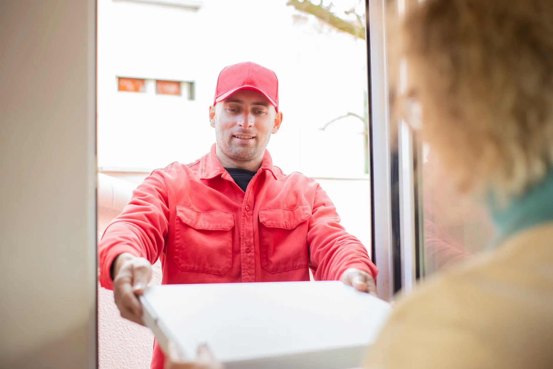 Repartidor con gorra y chaqueta roja entregando una caja a un cliente en la puerta de un edificio.