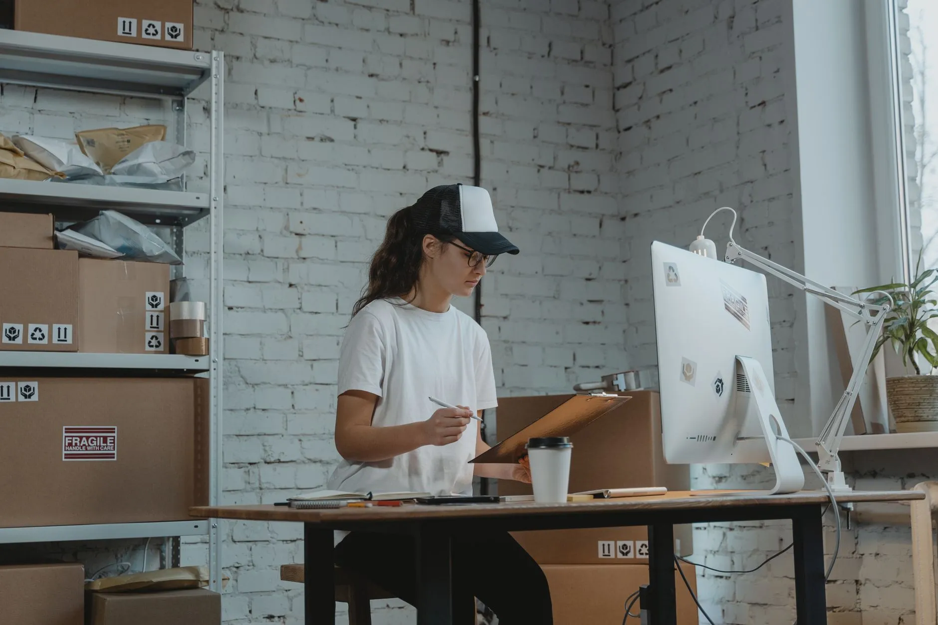 Mujer revisando un documento en una tablilla junto a un ordenador, en una oficina logística rodeada de cajas.